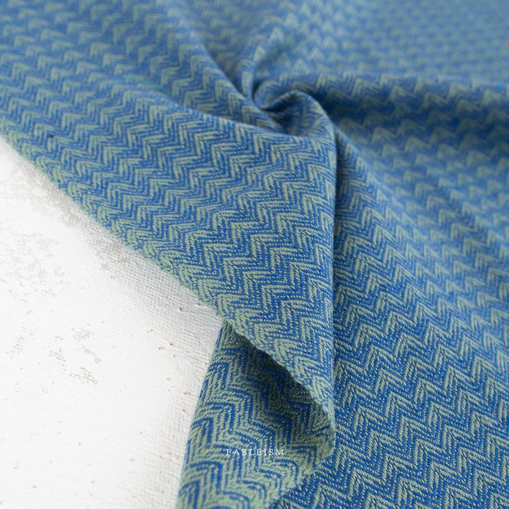 A close-up of blue fabric with a repeating light blue and white chevron or zigzag woven pattern, draped with a central fold on a light neutral background.