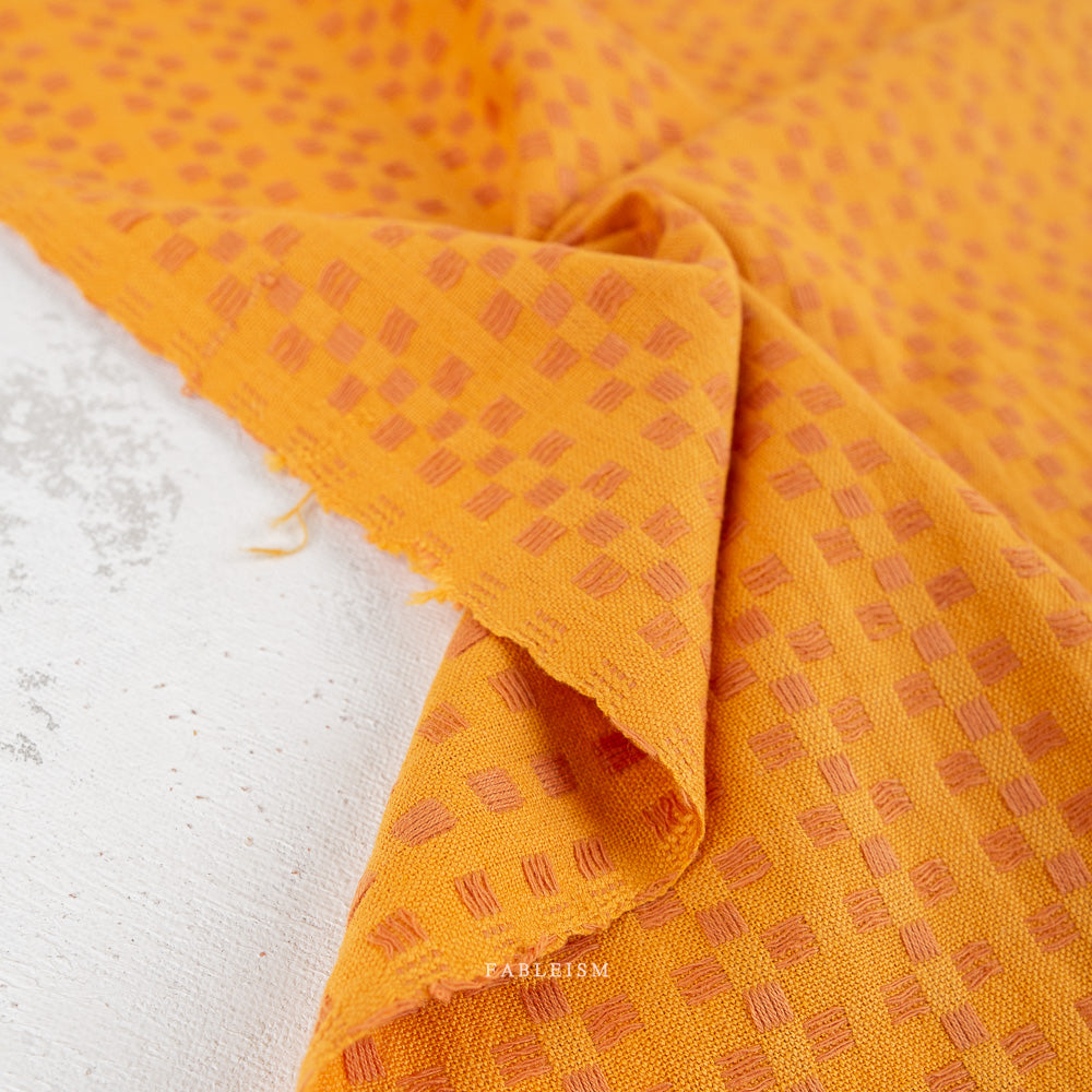 Bright orange fabric with a repeating small geometric pattern, draped to show texture and color, on a light neutral surface.