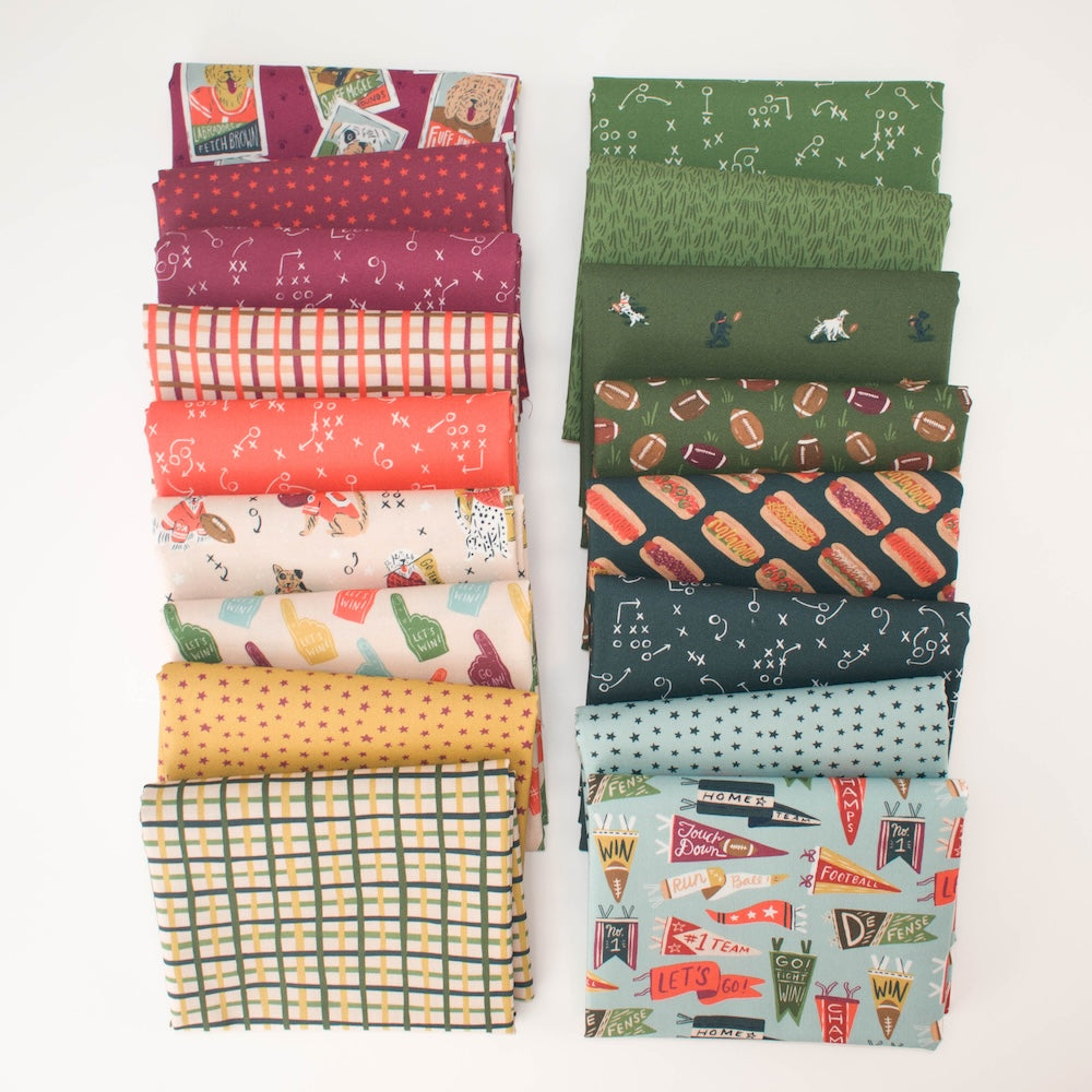 A tall stack of folded fabrics in assorted colors and dog football themed patterns, including greens, blues, reds, oranges, yellows, and whites, photographed on a clean white background.